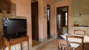A cozy room with a television on a wooden stand, a dining table with chairs, and a kitchen area with cabinets and a tiled backsplash. - NOBLE FARM HOLIDAY APARTMENT (Montepulciano Siena)