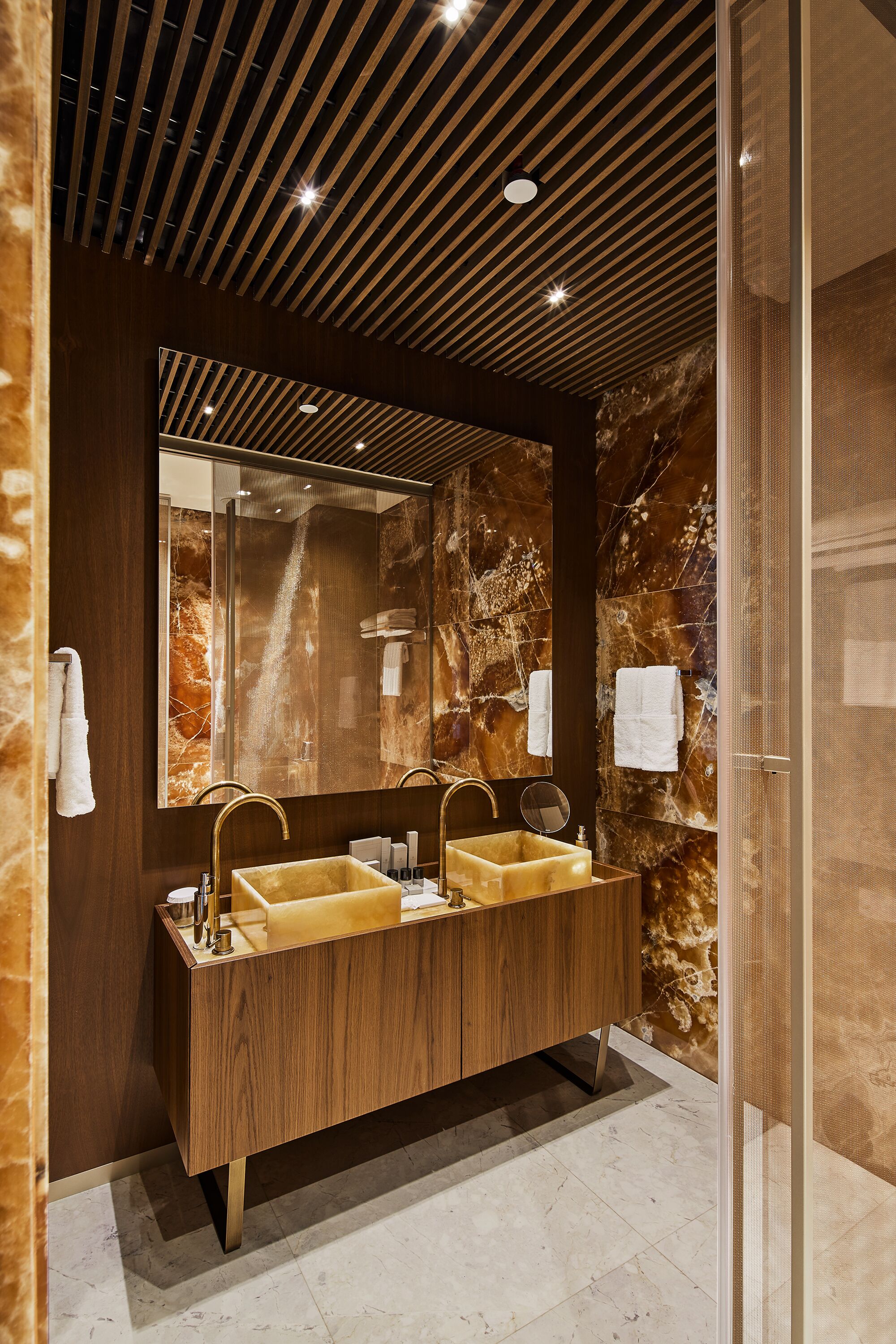 A modern bathroom with a wooden vanity, two sinks, a large mirror, and marble walls.