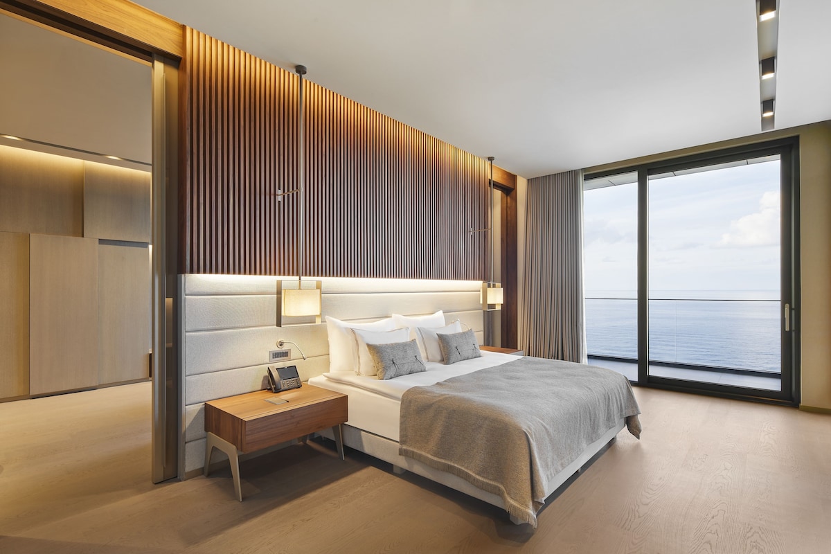 A modern hotel room with a large bed, bedside tables, a desk, and a view of the sea.