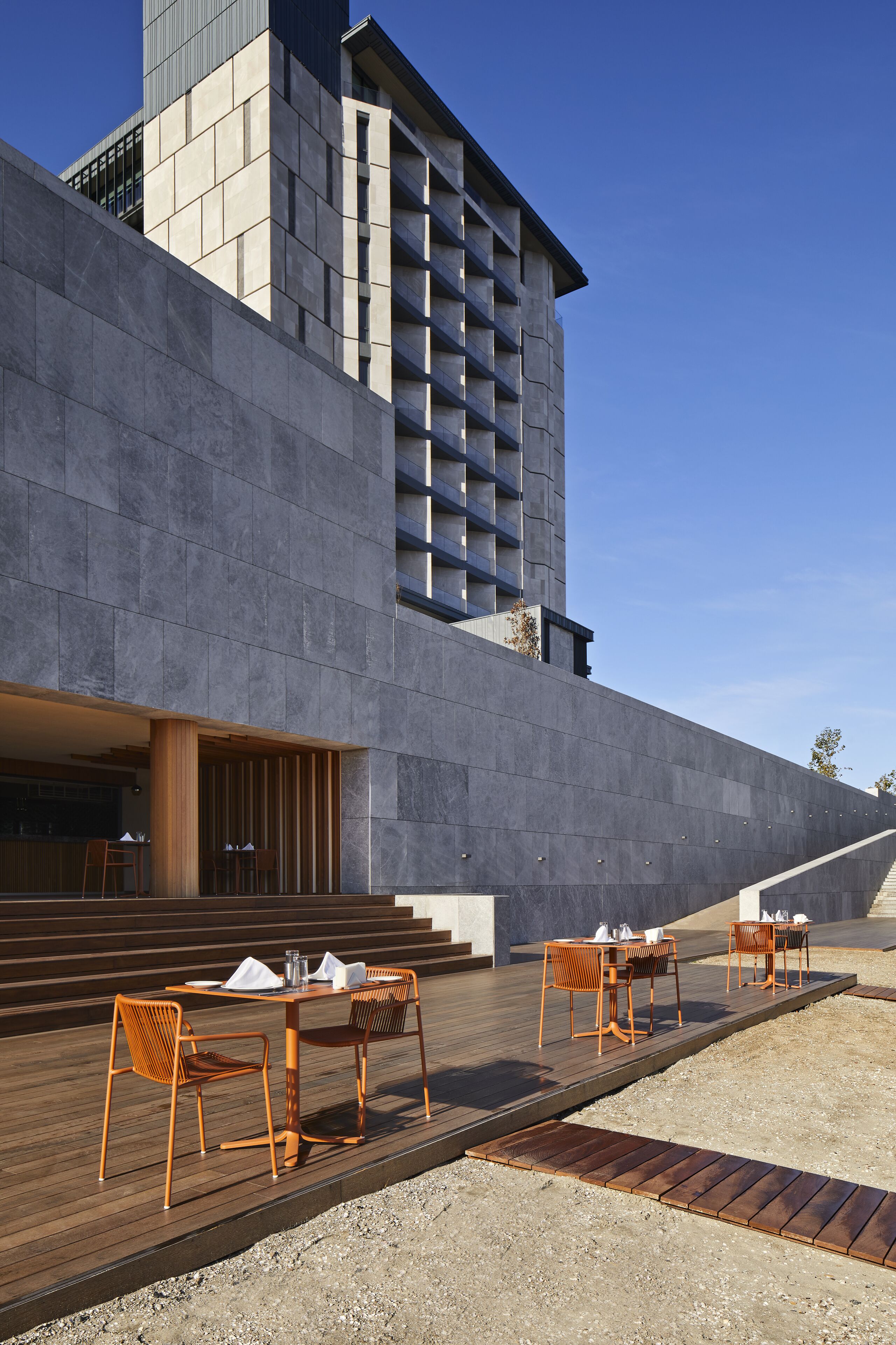 Un edificio moderno con fachada de hormigón, zona de estar al aire libre con mesas y sillas de madera, y una escalera que conduce a un nivel superior.