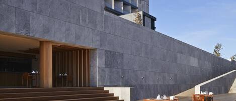 Un edificio moderno con fachada de hormigón, zona de estar al aire libre con mesas y sillas de madera, y una escalera que conduce a un nivel superior.