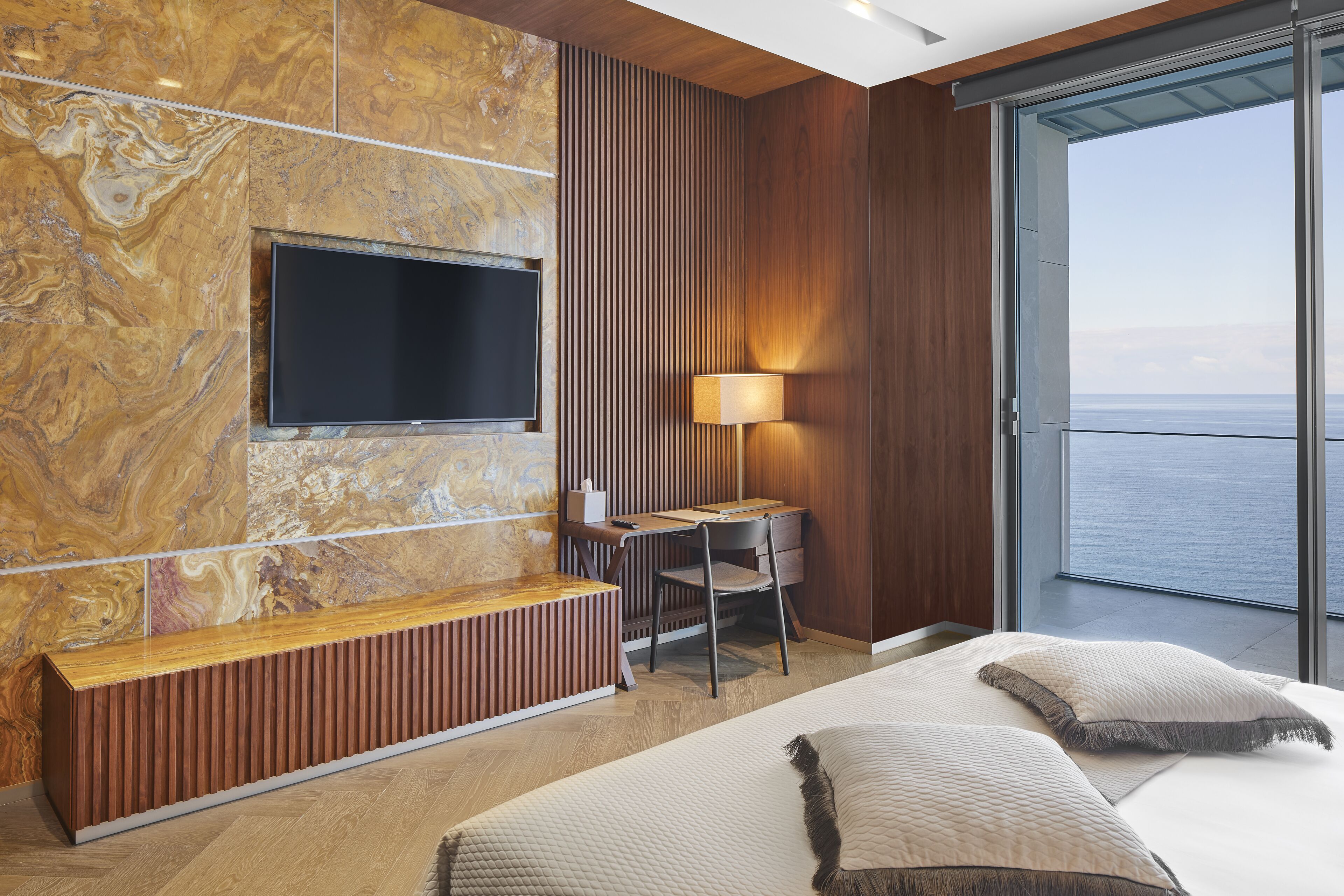 Una habitación de hotel moderna con un televisor grande, un escritorio con silla, una mesita de noche y una cama con almohadas.