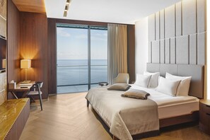 A modern hotel room with a large bed, a desk, and a view of the sea.