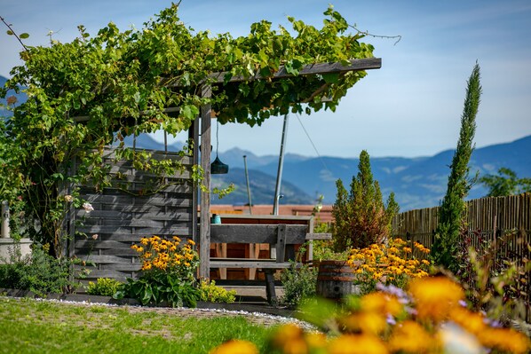 Een houten pergola met klimplanten, een bankje en een houten hek in een tuin met bloemen en een berg op de achtergrond.