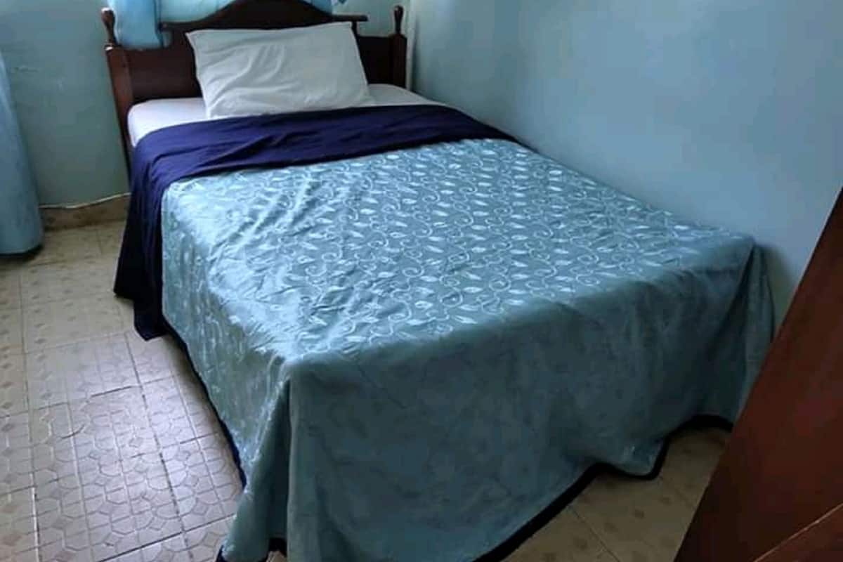 A single bed with a patterned bedspread, a wooden headboard, and a white pillow. The room has light blue walls and a tiled floor.