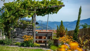 Een houten pergola met klimplanten, een bankje en een houten hek in een tuin met bloemen en een berg op de achtergrond.