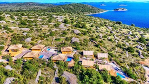 A resort with multiple thatched-roof cottages, each with a pool, set against a backdrop of a mountainous landscape and a clear blue sea. - Luxury Bungalow New Horizons IV (Murter)