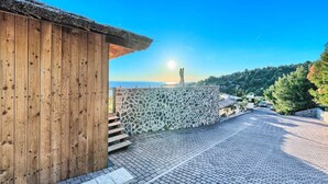 Edifici de fusta amb un mur de pedra i un carrer pavimentat que condueix a una vista del mar i una muntanya.
