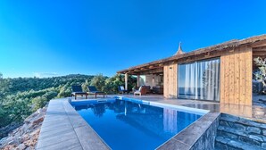 Ein moderner Poolbereich im Freien mit einer Holzterrasse, einer Steinmauer und Blick auf einen bewaldeten Hang.
