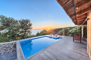 Piscina con vista al atardecer, terraza de madera y sillones de descanso.