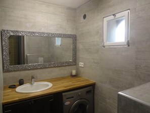 Un baño con tocador de madera, un lavamanos, un espejo con marco decorativo, una lavadora y una ventana.