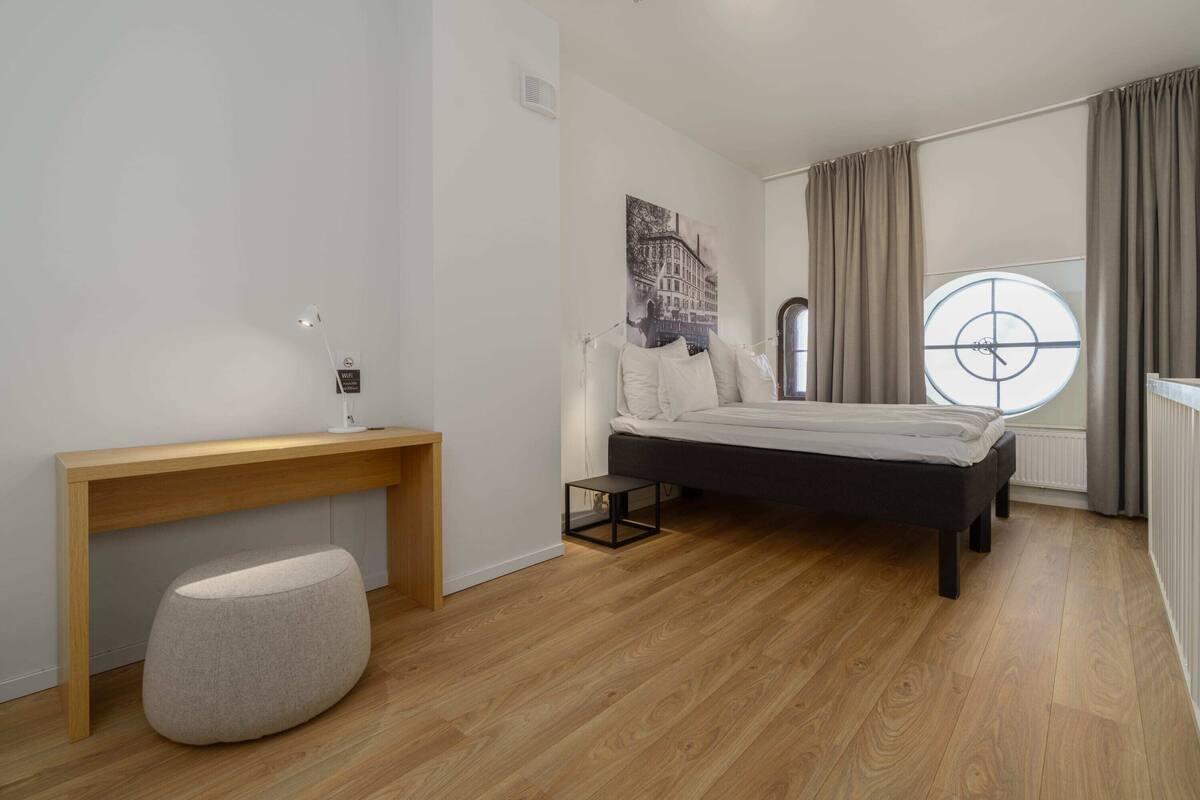 A bedroom with a bed, a desk, a chair, a window with curtains, and a wall-mounted light fixture.