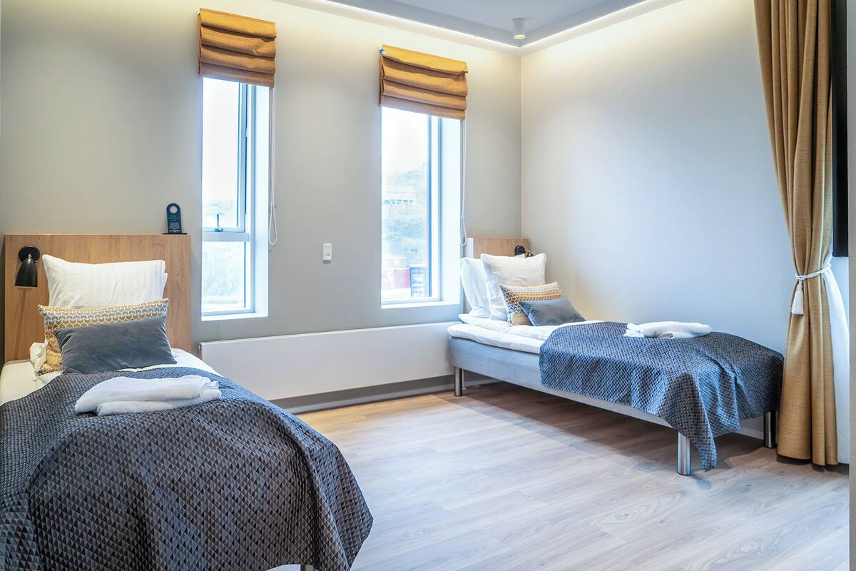 Een moderne hotelkamer met twee bedden, grote ramen met gordijnen, een houten hoofdbord en een zichtbaar stadsgezicht.