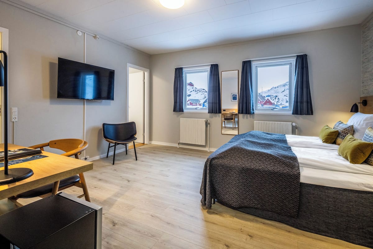 Een moderne hotelkamer met een houten vloer, een bed met een geborduurde deken, een bureau met stoel, een flatscreen televisie en een raam met uitzicht op een besneeuwde berg.
