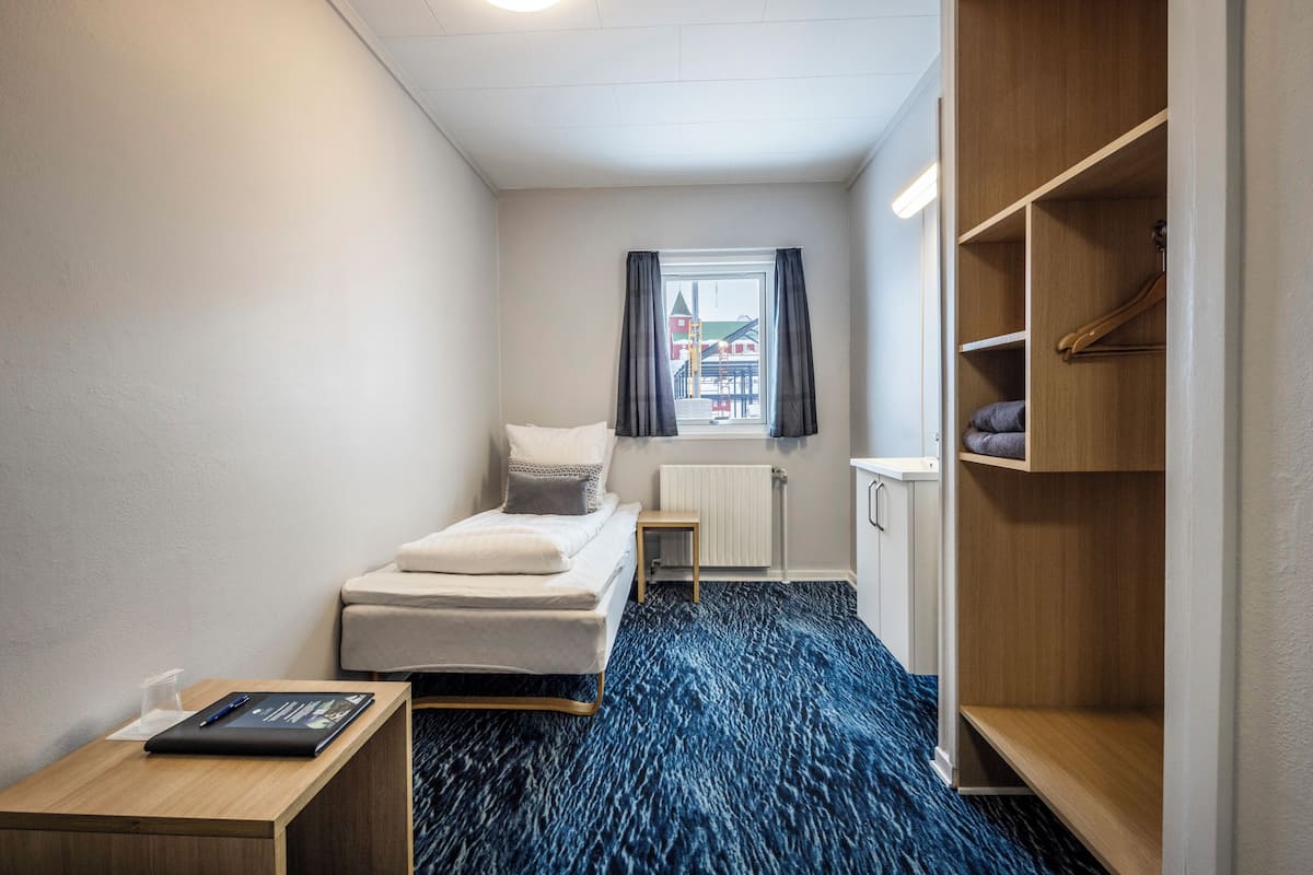 Een kleine, verzorgde hotelkamer met een bed, nachtkastje, kast en raam met gordijnen.