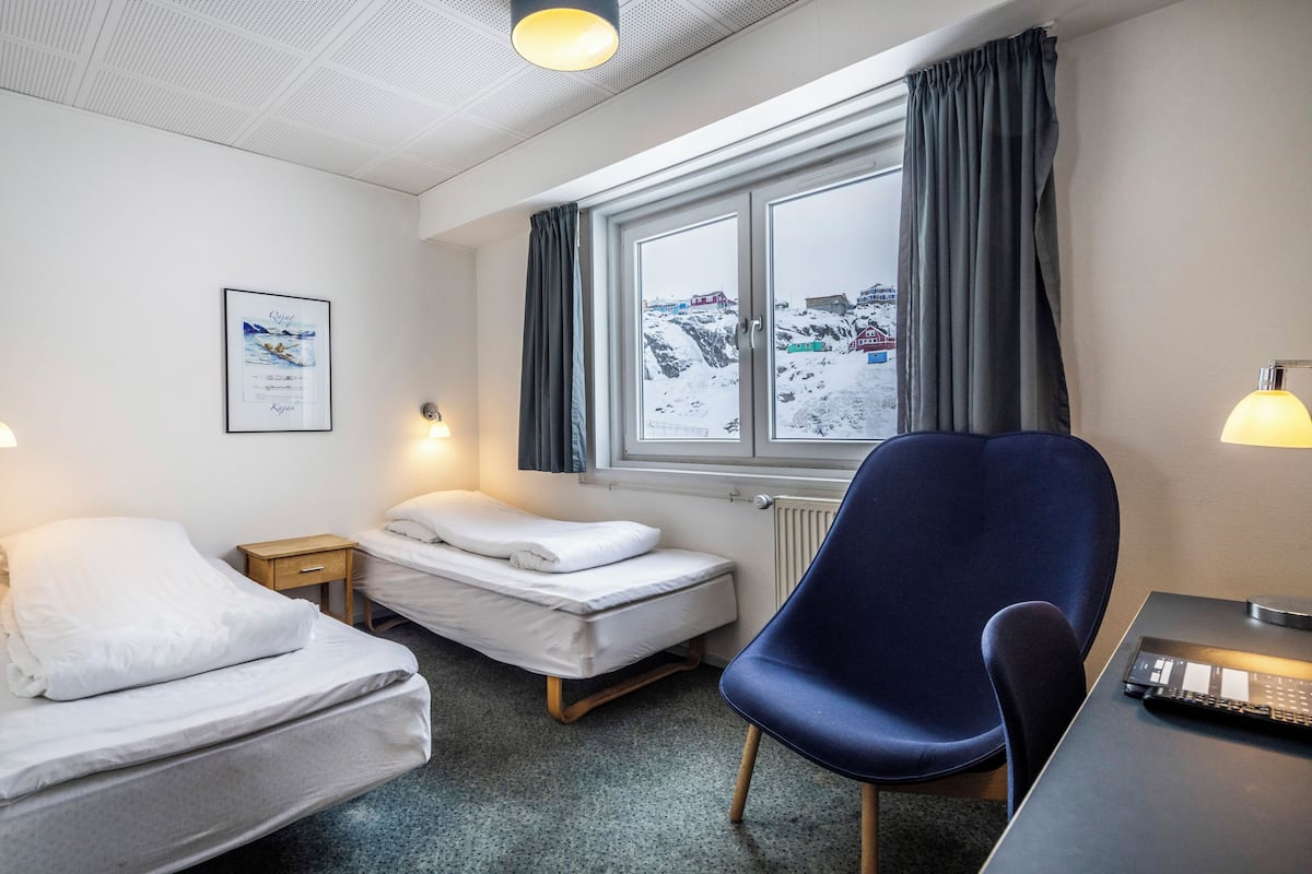Een hotelkamer met een bed, een bureau met een computer, een stoel, een raam met gordijnen, en een ingelijst schilderij aan de muur.