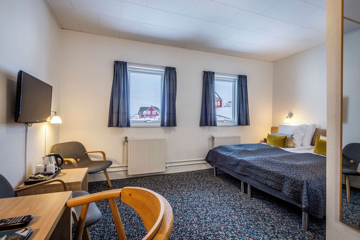 Een hotelkamer met een bed, twee ramen met gordijnen, een bureau met stoel, een televisie en een radiator.