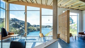 Ein geräumiges Zimmer mit großen Fenstern und Blick auf eine Küstenlandschaft. Es gibt mehrere Sitzbereiche mit Stühlen und Tischen.