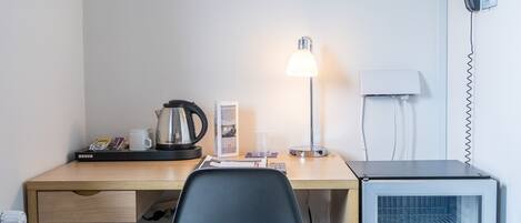 A compact hotel room with a wooden desk, a black office chair, a small refrigerator, a trash bin, a potted plant, and a wall-mounted phone.