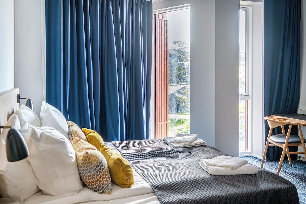 Hotelkamer met een bed, kussens, een bureau en uitzicht op een bergachtig landschap door het raam.