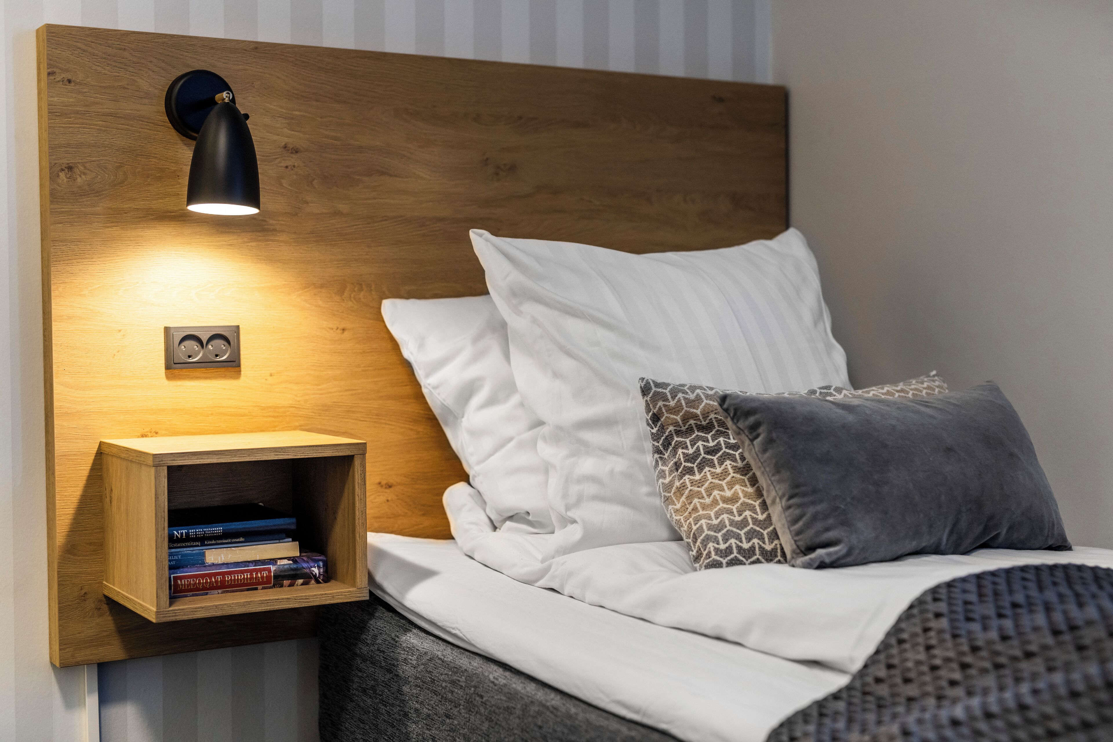 Una cama bien hecha con almohadas blancas y estampadas, una mesita de noche de madera con libros y una lámpara fijada a la pared.