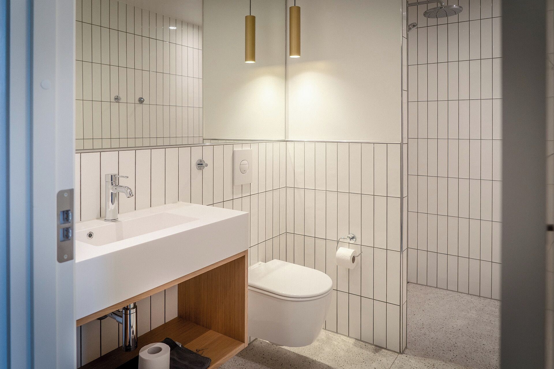 Un baño moderno con lavamanos, inodoro y ducha blancos. Hay dos lámparas colgantes sobre el lavamanos y un espejo grande sobre el inodoro.