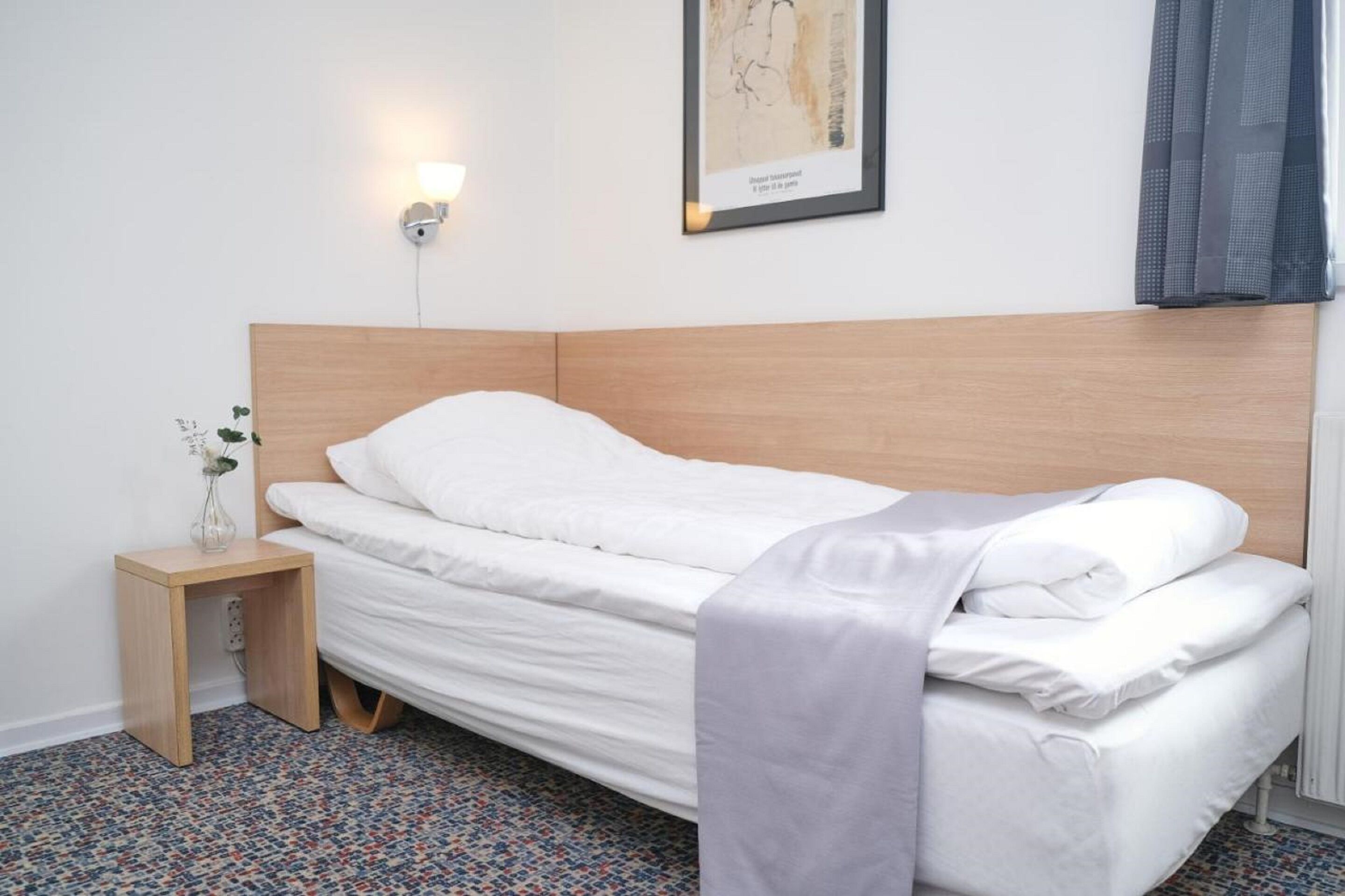 A neatly made bed with white linens, a small bedside table with a vase, and a wall-mounted light fixture.
