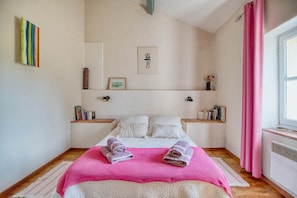 A neatly arranged bedroom with a bed, pillows, a pink blanket, and a bedside table with books.