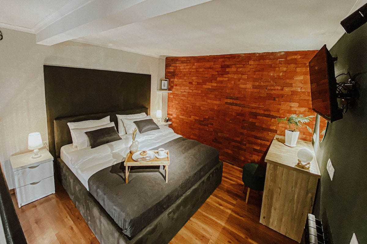 A modern hotel room with a large bed, a small desk, a brick accent wall, and a wooden floor.
