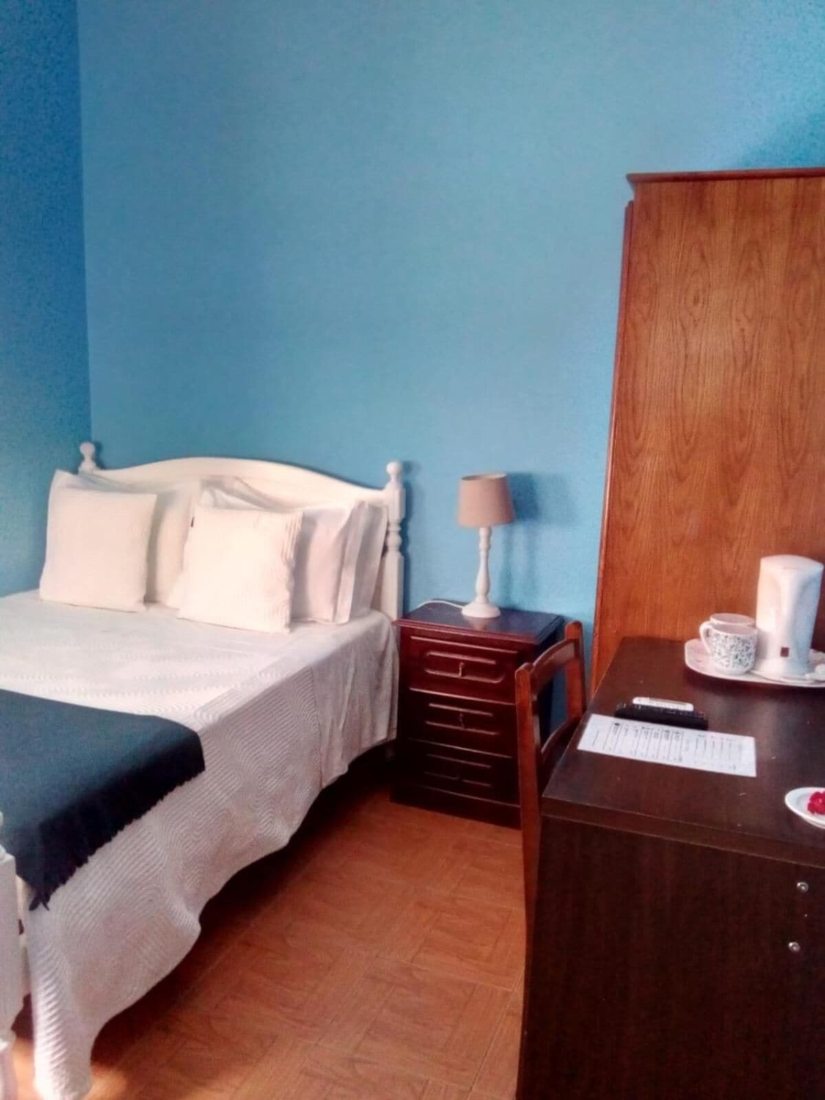 A hotel room with a bed, a nightstand with a lamp, a wooden cabinet, and a table with a cup and a remote control.