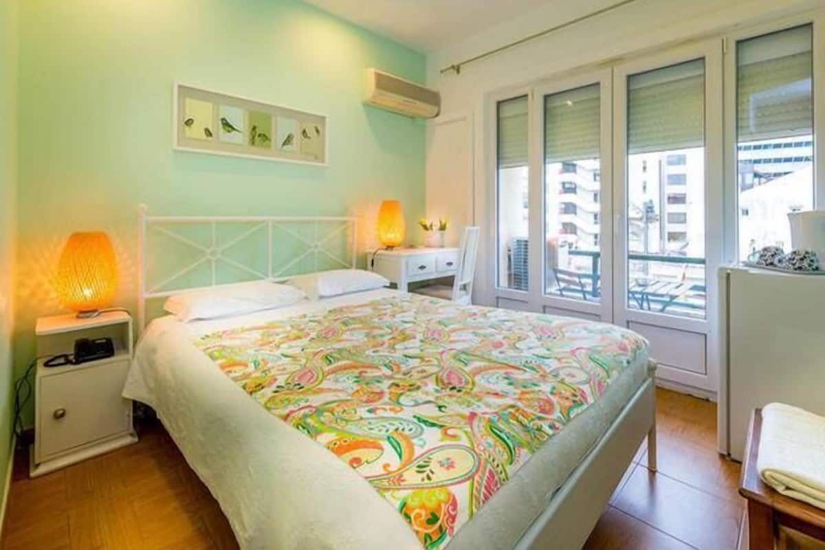 Un dormitorio con cama, lámparas de noche, cómoda y ventana con vistas a edificios.