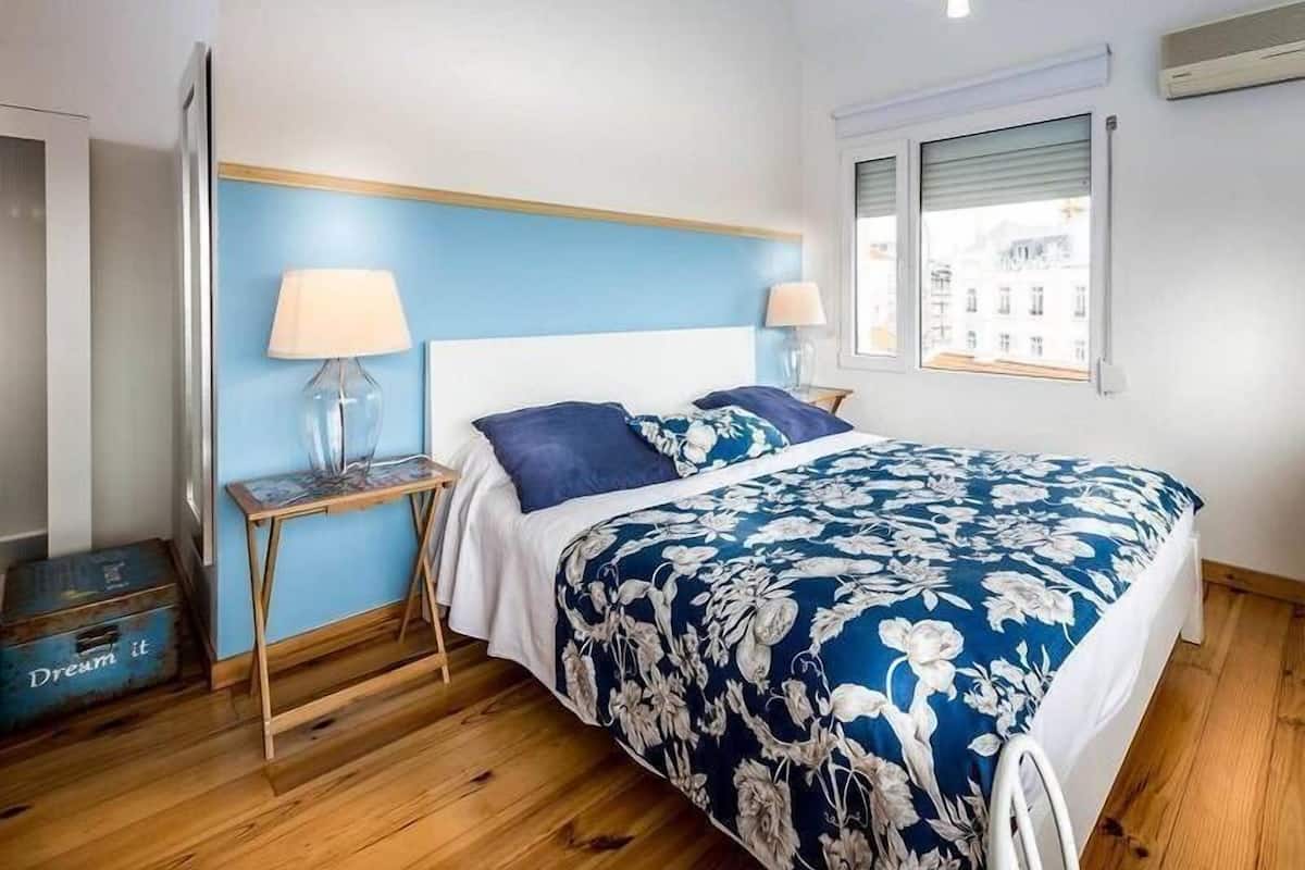 Un dormitorio con suelo de madera, una cama con colcha estampada, dos lámparas de noche, una mesita de noche y una ventana con persianas.