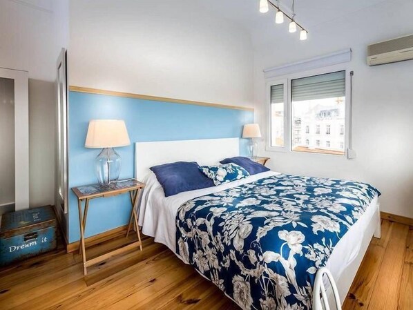 A bedroom with a wooden floor, a bed with a floral bedspread, two bedside lamps, a nightstand, and a window with blinds. - Casa Belmonte (Lisbon)