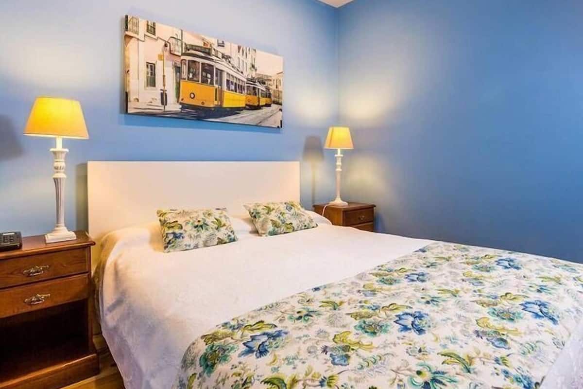 Un dormitorio con una cama, dos mesitas de noche, un cuadro de una escena callejera y una colcha estampada con motivos florales.