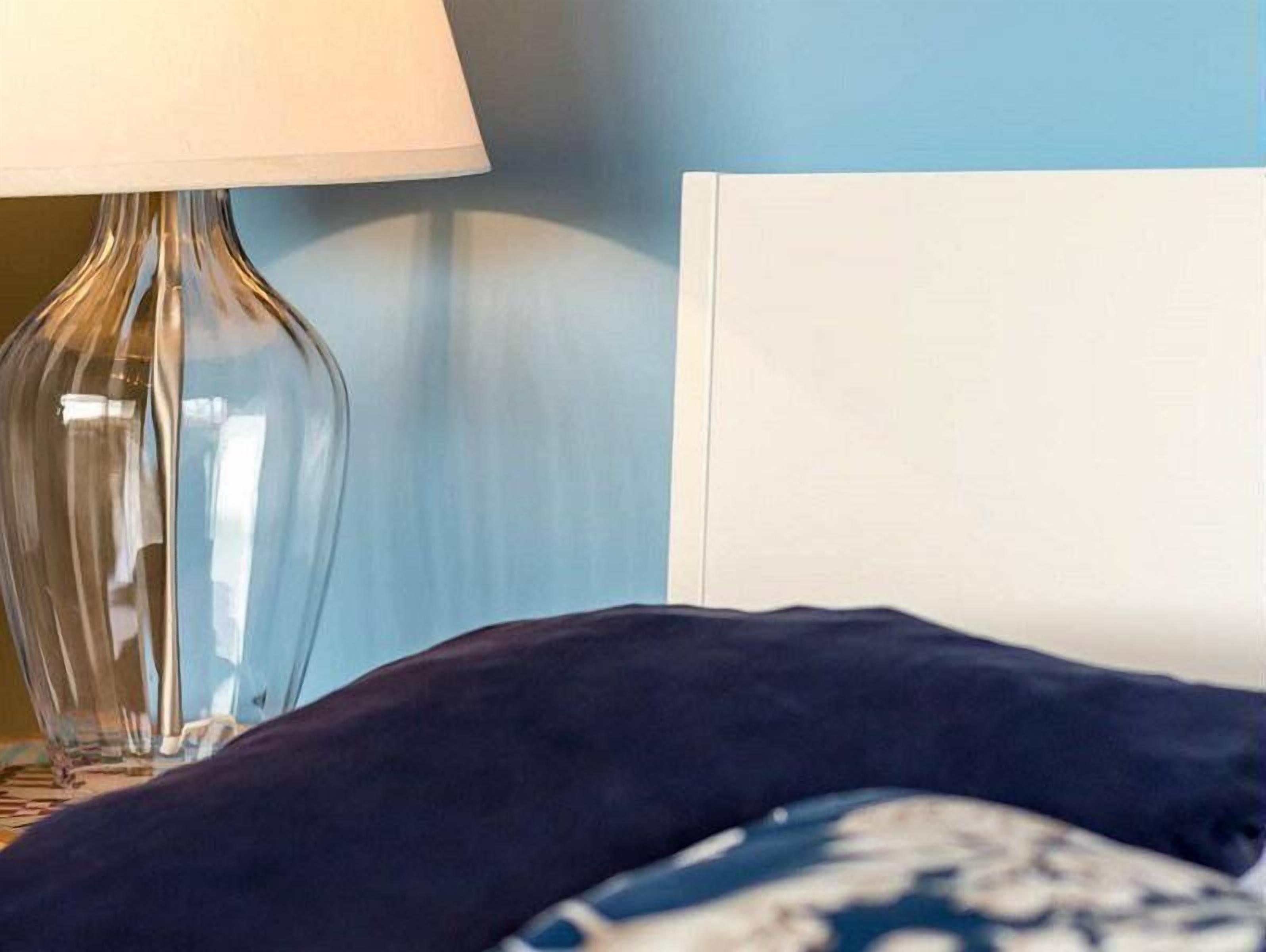 A bed with a dark blue headboard, a clear glass lamp, and a white wall in the background.