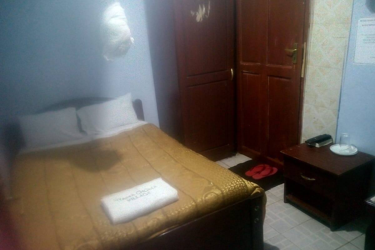 A hotel room with a bed, a wooden wardrobe, a bedside table with a phone and a glass, and a wall-mounted light fixture.