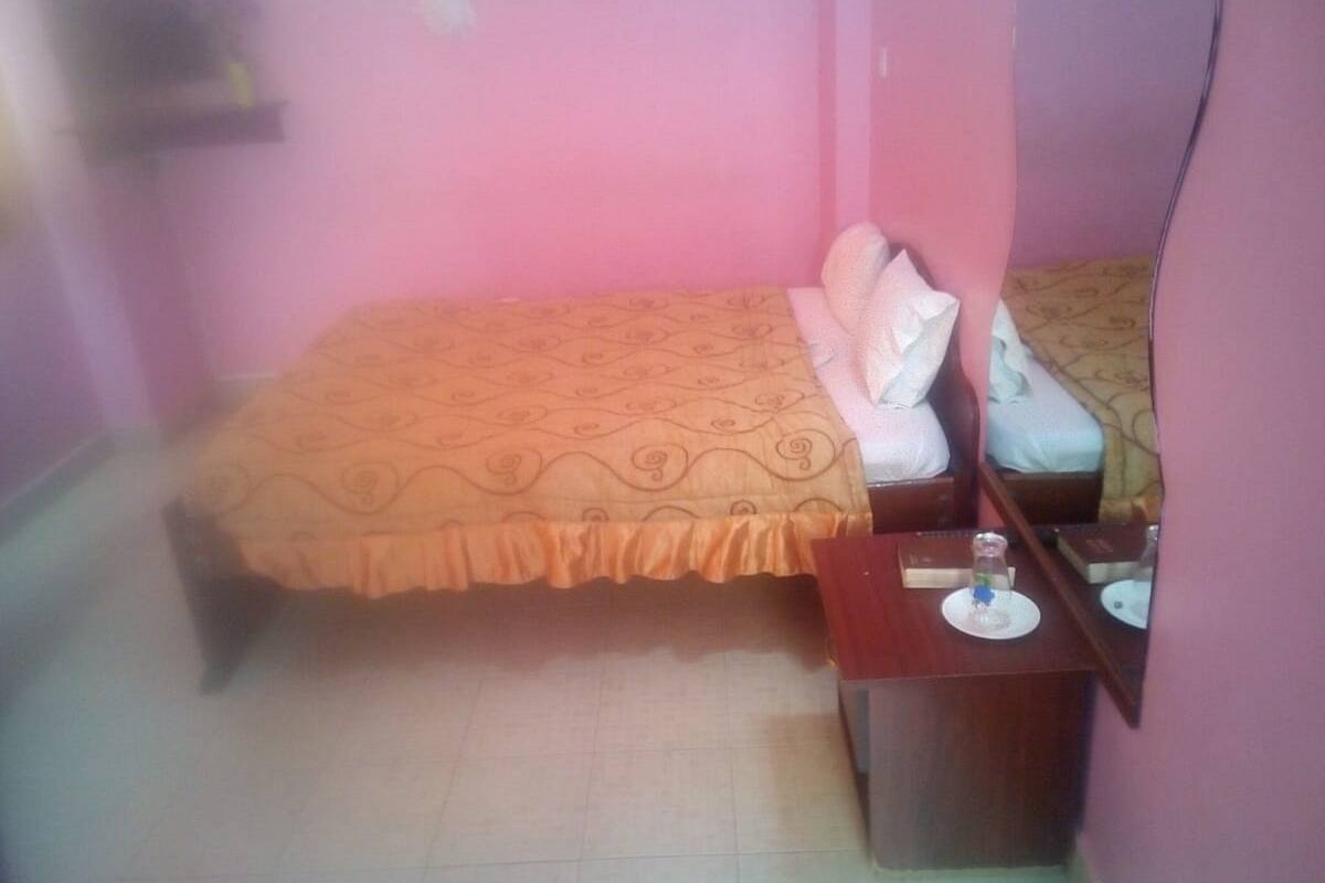 A small, pink-walled room with a bed, a bedside table, and a mirror.