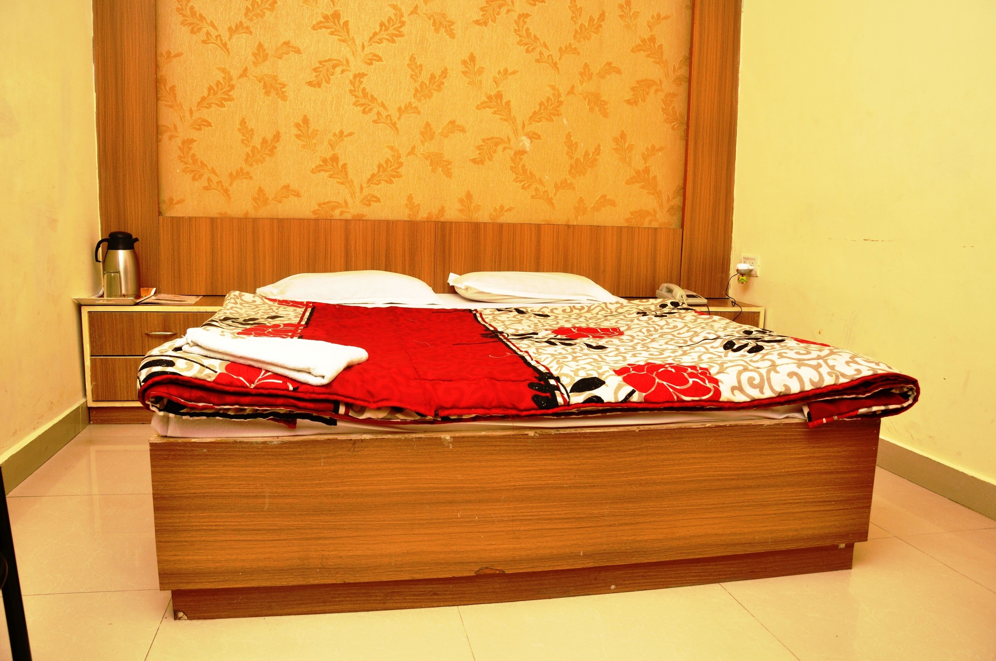 A neatly made bed with a red and white patterned blanket, a wooden headboard, and a nightstand with a kettle and a phone.