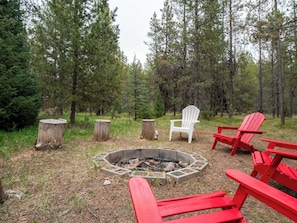 A campfire surrounded by red and white Adirondack chairs in a forest setting.