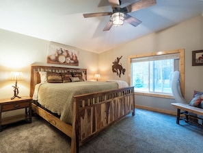A bedroom with a wooden bed, two bedside tables with lamps, a ceiling fan, a window with a view of trees, and a wall decoration of a cowboy.