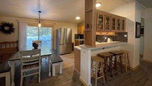 A kitchen with wooden cabinets, a stainless steel refrigerator, a dining table with chairs, and a bar area with stools. - Wilderness Pines, Close to Yellowstone, Sleeps 10 (Island Park)