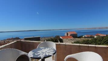 Una terraza con sillas de mimbre y una mesa, con vistas a una zona costera con casas y un cielo azul despejado.