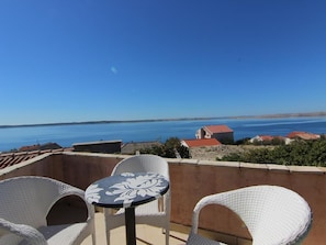 A terrace with wicker chairs and a table, overlooking a coastal area with houses and a clear blue sky.