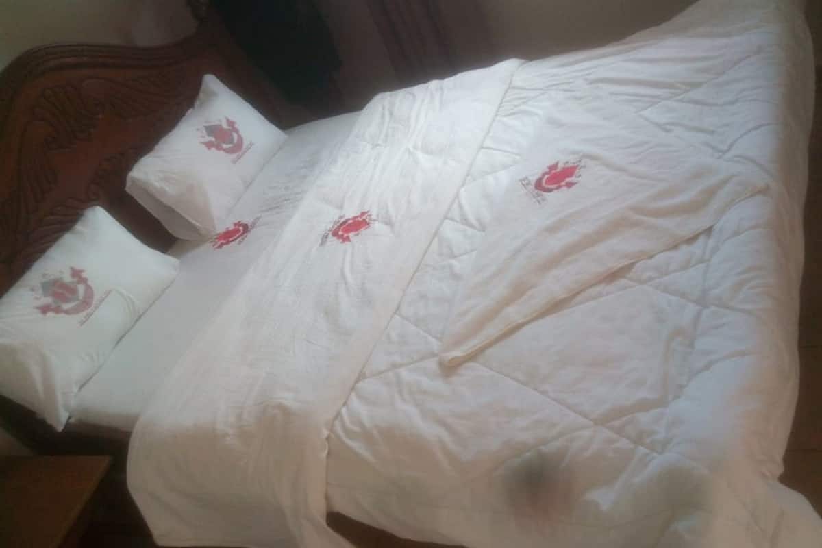 Una cama con colcha blanca y almohadas, cabecera de madera y suelo de baldosas.