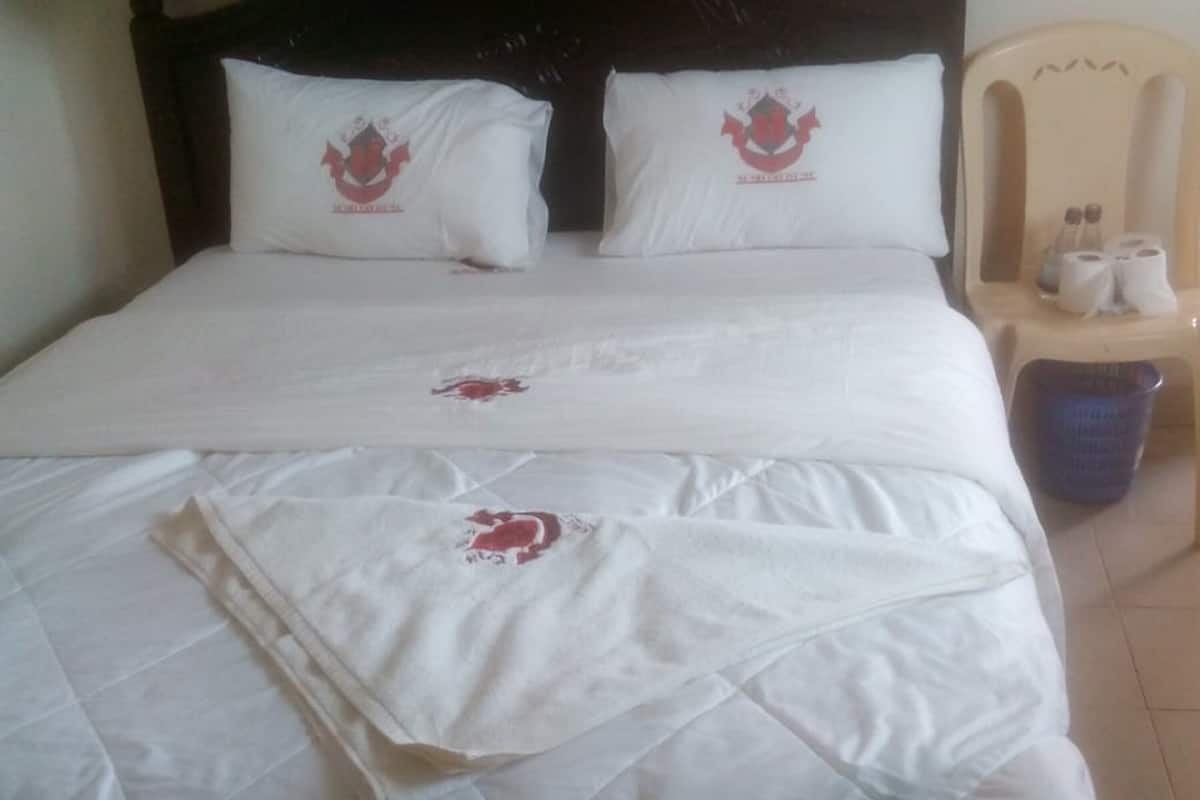 Una cama con ropa blanca y dos almohadas, una silla con artículos de aseo, y un cubo de basura.