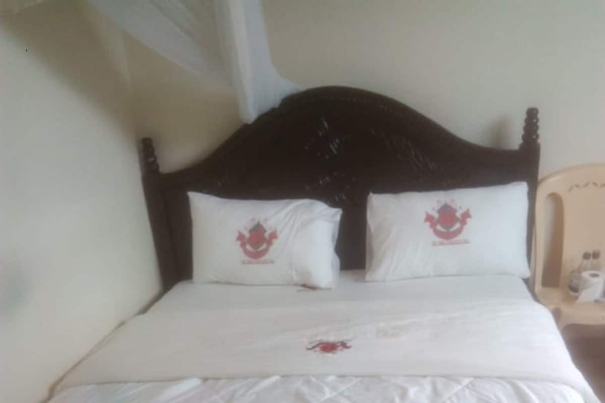 Una cama con una colcha blanca acolchada, dos almohadas con un escudo rojo y un cabecero de madera.