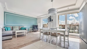 Un salon moderne comprenant un canapé, une table basse, une table à manger et des chaises. Une grande fenêtre offre une vue sur le ciel et les bâtiments.