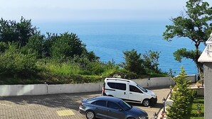 A parking area with two cars, a white van, and a grey sedan. The background features a clear blue sky and a view of the sea. - Amasra Sunrise Otel (Amasra)