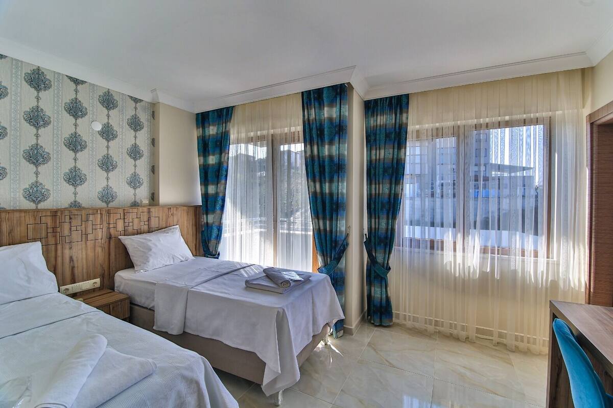 Habitación de hotel con dos camas, cabecero de madera, ventana grande con cortinas translúcidas y papel tapiz estampado.