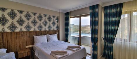 A hotel room with a large bed, a wooden headboard, a nightstand, and a view of the sea through a balcony.